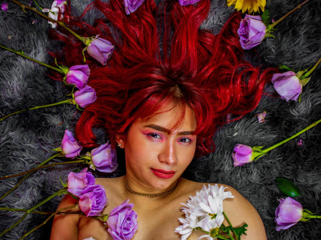 MelannyTaylorr with bright red hair, purple eye makeup, lying on a grey furry surface, surrounded by purple roses and a white daisy, holding flowers, wearing a gold necklace.