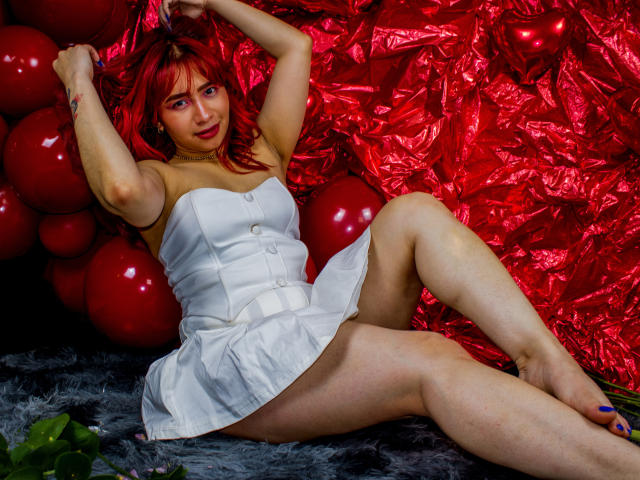 MelannyTaylorr with red hair, wearing a white dress, has her arms raised, showing her tits, and is lying down with her legs spread, exposing her pussy. Red balloons.