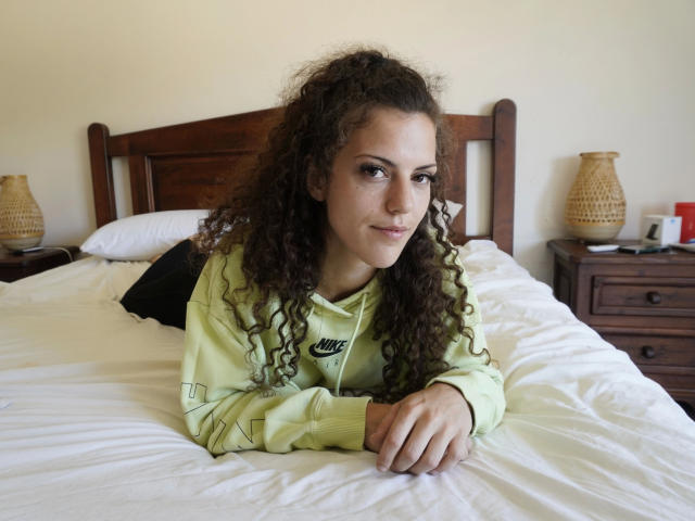 OliviaOcean-hot with curly brown hair, green Nike hoodie, lying on white bed, hands under chin, smiling subtly, wooden headboard, nightstands with baskets and red item, casual bedroom.