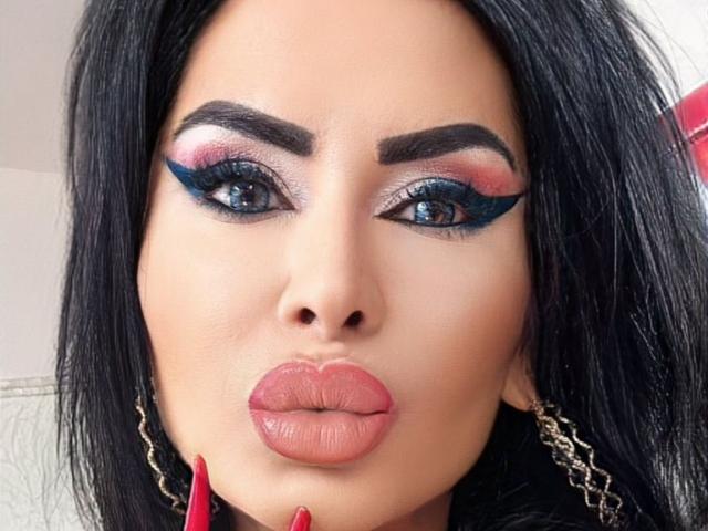 GoddessNaomi has blue eye makeup with black wings, pink and red eyeshadow, full pink lips, and black hair. Her eyebrows are thick and she's wearing red earrings.