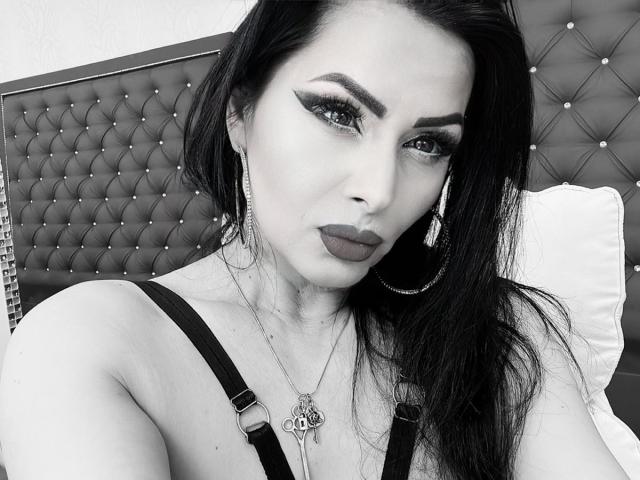 GoddessNaomi with dark, wavy hair, bold eyeliner, and dark lipstick, wearing a strap dress, hoop earrings, and a necklace, with a tufted headboard in the background.