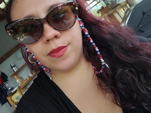 LexyDom with red lipstick, dark wavy hair, tortoiseshell sunglasses, and colorful beaded earrings, wearing a black top, smiling slightly, reflected in sunglasses.