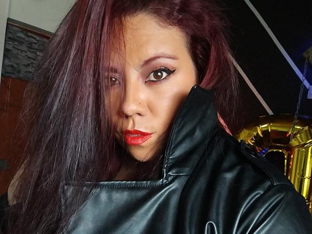 LexyDom with dark red hair, black leather jacket, red lipstick, and winged eyeliner, looking seductively at the camera, her ass barely visible under the jacket.