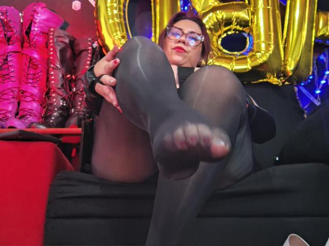 LexyDom with glasses, red lipstick, black sheer tights, and black dress, showing her foot, with her pussy slightly visible through tights, sitting on a black couch.