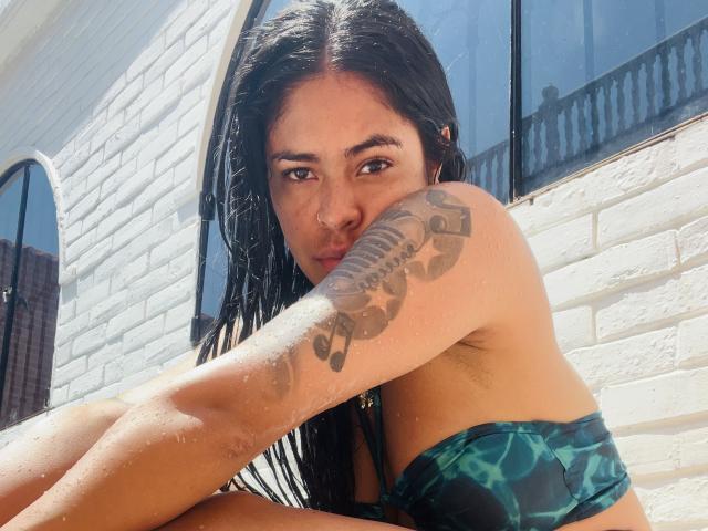 StellaBeckker met nat zwart haar, een groene en zwarte tie-dye bikini, een tatoeage op haar rechterarm, zittend buiten, met zonlicht op een witte bakstenen muur, en een zwart raam op de achtergrond..