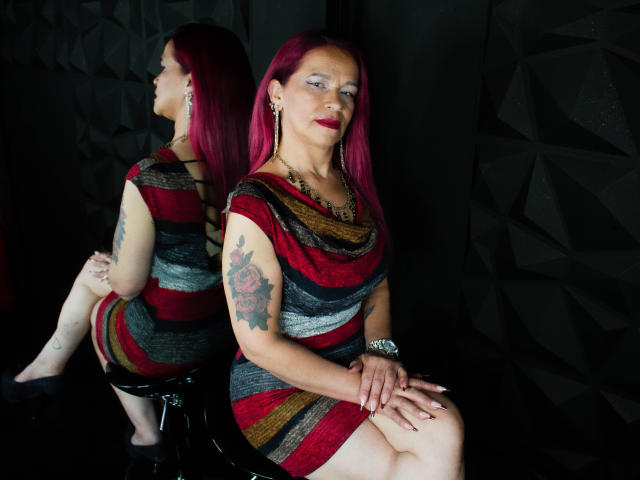 LizhCarthner with pink hair, red dress, striped pattern, rose tattoo, sitting, hands on knees, black wall, reflection, red lipstick, earrings, watch, black chair, confident, sexy.