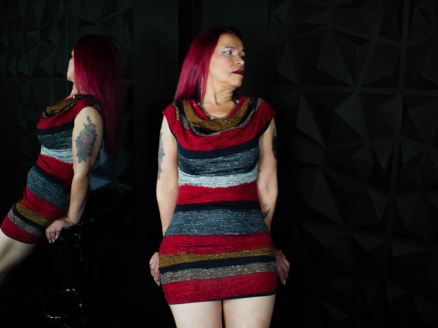 LizhCarthner with red hair, striped dress, tattoos, and pale skin, leaning against a black chair, reflected in a mirror, showing her ass and small tits.