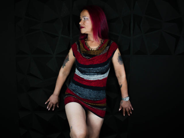 LizhCarthner with red hair, striped dress, tattoos on arms, red lipstick, silver watch, standing against dark geometric wall, hands on sides, looking to the right.