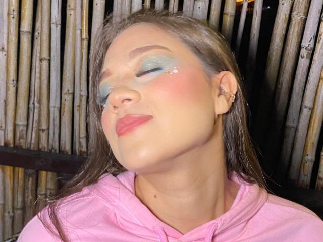 LindaJimenex with closed eyes, wearing pink hoodie, has blue-green eyeshadow, pink blush, and glossy pink lips. Bamboo sticks in background. She's sporting small earrings and wet brown hair.