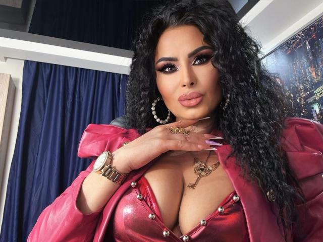 GoddessNaomi with curly black hair, heavy makeup, and pink lipstick, wearing a red leather jacket, revealing her big boobs in a shiny red top with gold studs.