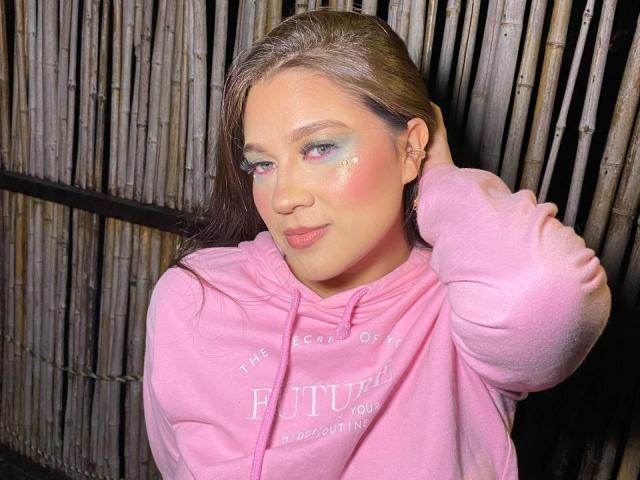 LindaJimenex with brown hair, pink hoodie, blue-green eyeshadow, glitter on cheeks, hand in hair, subtle smile, pink lips, bamboo wall in background, medium shot, casual style.