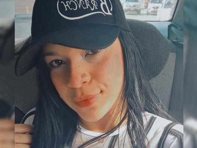 SteefyCourny with black hair, wearing a black cap and striped shirt, winking, nose ring, and red lipstick, sitting in a car, holding something with a blurred hand.