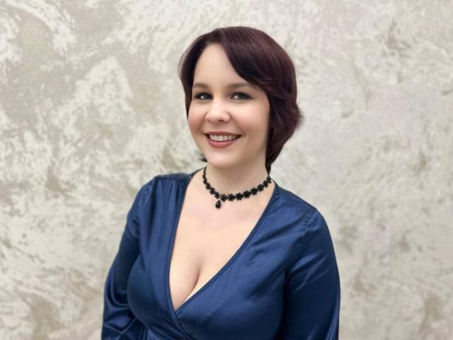 JasmineParadize, con i capelli corti e scuri, di colore rosso, sorride, indossando un abito di seta blu con un profondo scollo, una collana di perline nere, e mostrando il seno. È carina e sexy..