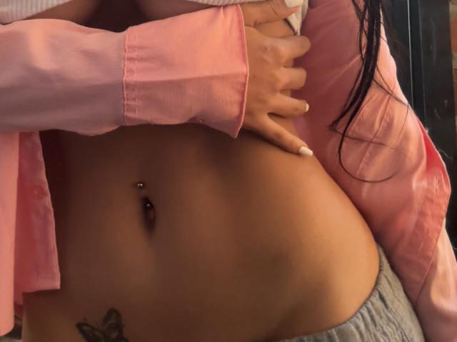 SophiePaytton's bare, tan-skinned belly with a navel piercing and a tattoo below her belly button, wearing a pink shirt and gray pants, has her hand on her chest.