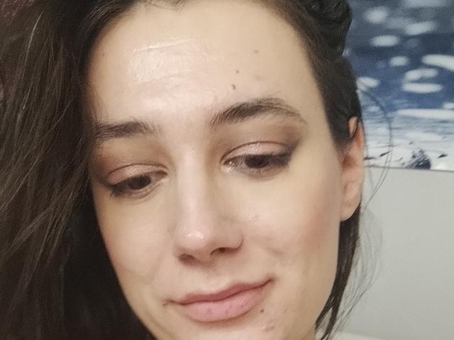 JuliaHuntis with a slightly shiny forehead, brown eyes, subtle eyeliner, dark brown hair, and a small, gentle smile, showing a few small blemishes on her face.