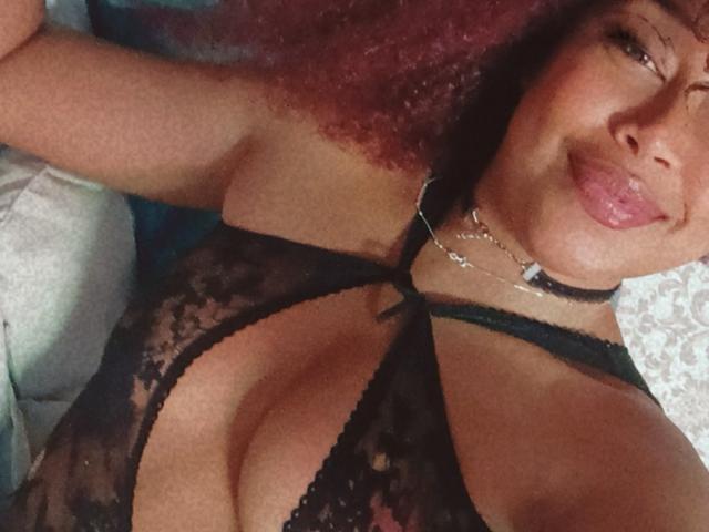 DalilaHeinz with curly red hair, tongue out, wearing a black lace bra that exposes her big boobs, and a black choker with a silver necklace.