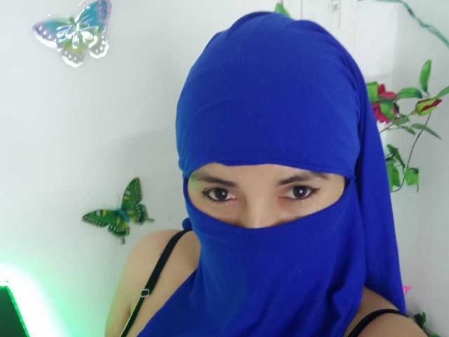 TrishaXx usando um lenço azul que cobre o nariz e a boca, com os olhos castanhos visíveis, uma blusa com alças pretas e borboletas azuis na parede branca..