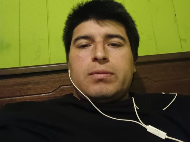 HotChileGuy with short black hair, white earbuds, and a black shirt, looking slightly bored or tired, face centered, green wall in background.