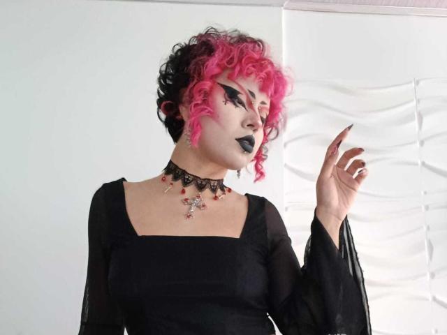 GothVioleth tiene el pelo rizado de color rosa, maquillaje negro y lápiz labial gótico. Lleva un vestido negro, un collar con gemas rojas y tiene una mano levantada. Tiene la piel clara y los ojos negros..
