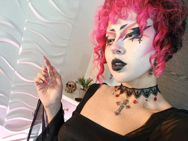 GothVioleth met roze krullend haar, witte schmink, zwarte lippenstift, een zwarte jurk, een kant kraagje, een rode kruis hanger en zwarte nagellak, houdt een vinger omhoog en kijkt naar de camera..