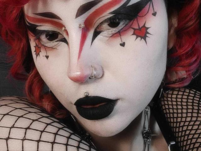 GothVioleth met witte make-up op het gezicht, rode en zwarte oogmake-up, zwarte lipstick, rood krullend haar, een neusring, een lipring en netruitmouwen, met een intense blik. De borsten zijn gedeeltelijk zichtbaar..