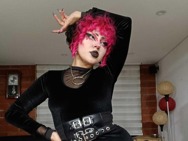 GothVioleth met roze krullend haar, een zwarte fluwelen jurk, zwarte lipstick en meerdere riemen, poseert met één arm omhoog, in een kamer met bakstenen muren en witte jaloezieën..