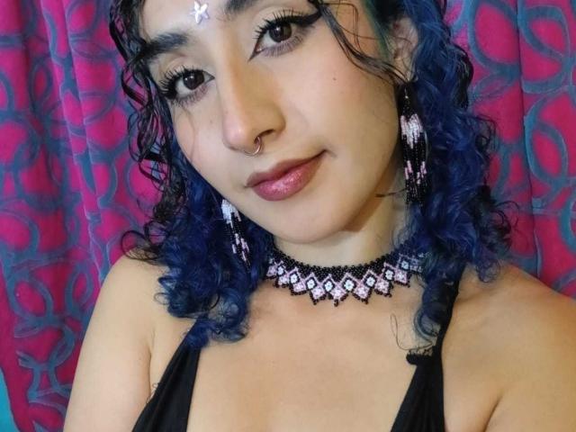 ValerieMasse has blue curly hair, a nose ring, and pink lipstick. She wears black earrings, a diamond choker, and a butterfly forehead accessory. Her tits are small.