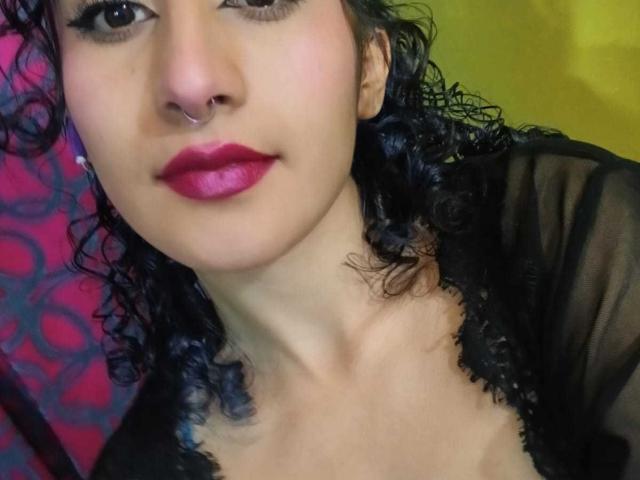 ValerieMasse tiene el pelo negro y rizado, un piercing en la nariz y un lápiz labial rojo brillante. Lleva una blusa negra transparente que deja ver parte de su escote. Sus ojos están ligeramente abiertos, mirando a la cámara..