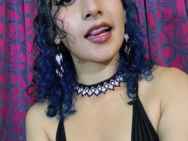 ValerieMasse, avec ses cheveux bouclés bleus, son anneau de nez et son haut de maillot noir à encolure plongeante, porte un collier de diamants et des boucles d'oreilles pendantes, avec des lèvres roses brillantes légèrement entrouvertes..