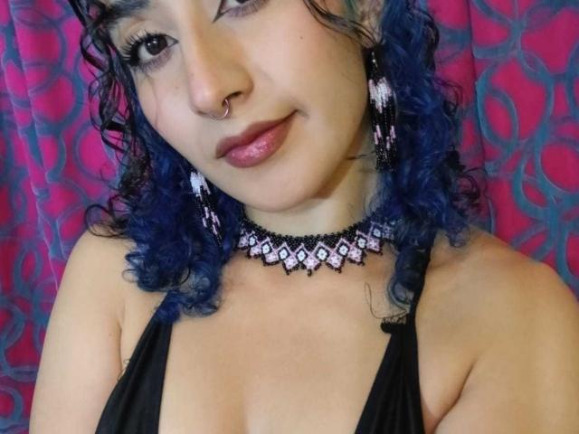 Valerie Masse hat blau gelocktes Haar, einen Nasenring, pinken Lippenstift, ein schwarzes Halstuch-Top, eine Diamantkette und pinke Diamant-Ohrringe. Ihre Brüste sind klein, und sie lächelt leicht..