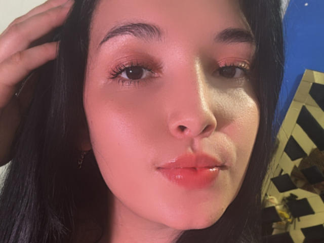 SarahGood's face, shiny pink lips, small nose, dark eyebrows, long black hair, hand in hair, slight pout, brown eyes, light skin, subtle makeup, glossy skin, slight smile.