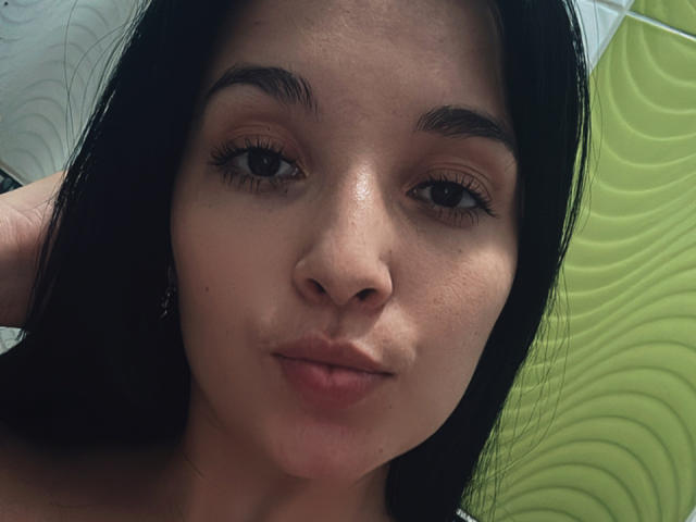 Le visage de SarahGood, cheveux foncés, yeux marron, lèvres pulpeuses, léger sourire, maquillage discret, petit nez, peau lisse, quelques taches de rousseur, elle regarde l'appareil photo avec une expression détendue..