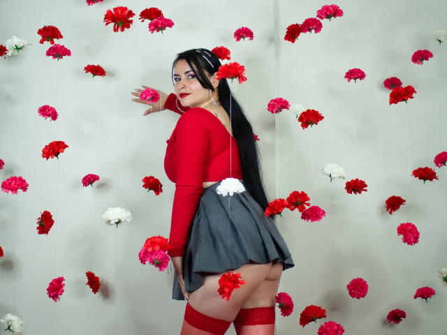 NicoleJons with long black hair, red top, gray skirt, red thigh-highs, and visible ass, surrounded by red and white flowers, looking back with red lipstick and white flower.