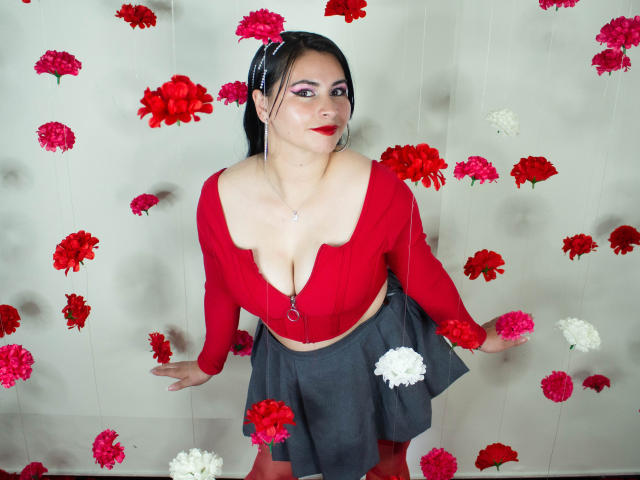 NicoleJons with big tits, red crop top, gray skirt, and red lipstick, holding flowers, black hair in a ponytail, pink eyeshadow, white wall with red and white flowers.