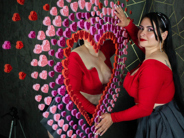 NicoleJons with black hair, red crop top, and black skirt, making a heart shape with pink and red floral petals, revealing her tits, against a dark background.