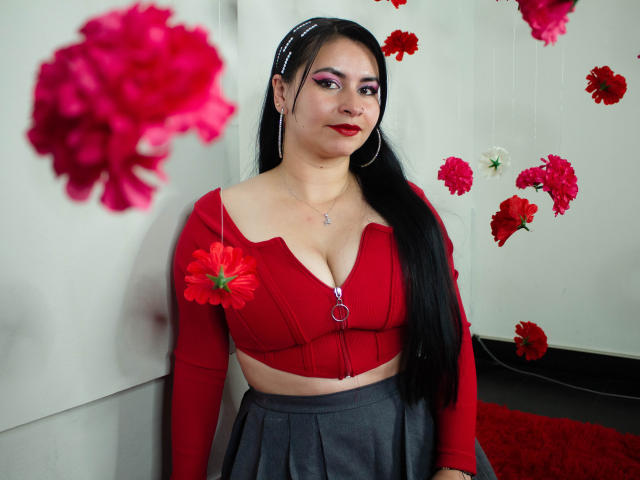NicoleJons has long black hair, red crop top showing her tits, gray skirt, red flower in hair, and hoop earrings, with pink and red flowers in the blurred foreground.