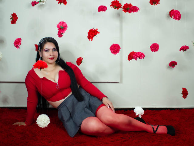 NicoleJons with long black hair, red crop top, gray skirt, red tights, black shoes, sitting on red carpet, surrounded by red and white flowers, holding a red flower.