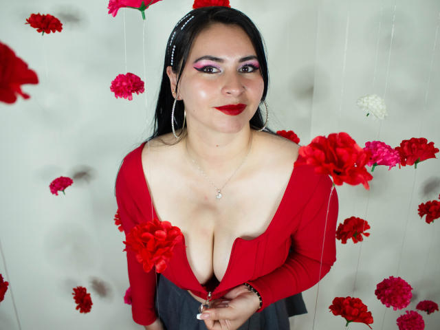 NicoleJons with red dress, low cleavage, large tits, pink eyeshadow, red lipstick, black hair, silver necklace, and red flowers in front and around her.