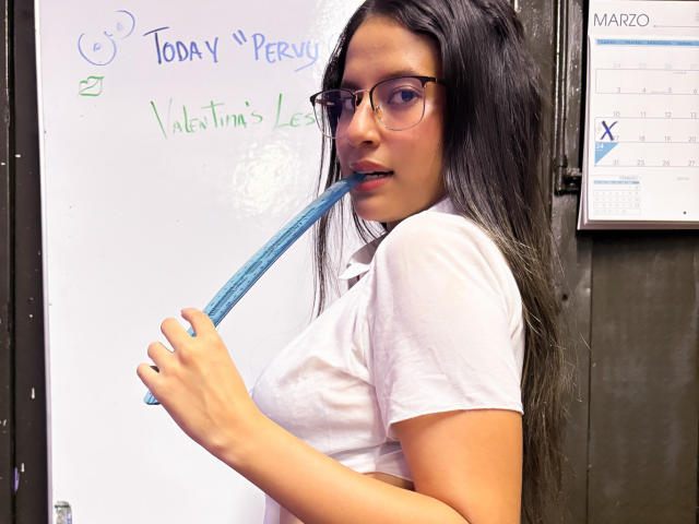 EroticValen with long black hair, glasses, white shirt, biting blue marker, holding it in her mouth, looking at camera, in classroom setting. Her tits are visible under shirt.