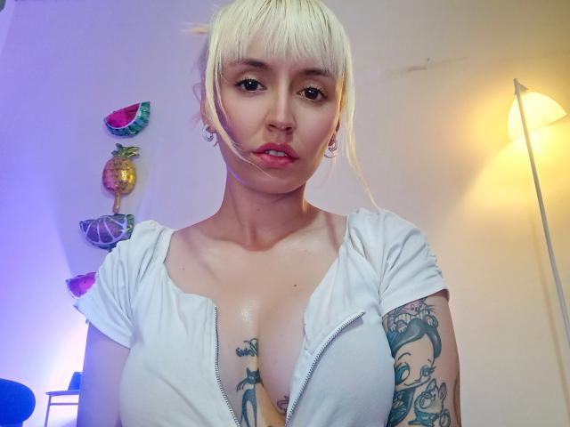 SaraleePoison with blonde hair, white zip-up top, visible boobs, tattoos on chest and arm, ear gauges, standing, looking at camera, pink lips, indoor lighting, wall art in background.