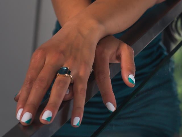 EvelinLuna's hands with teal and white nail polish, wearing a green ring and a gold ring, resting on a glass railing. Her fingers are slightly curled.