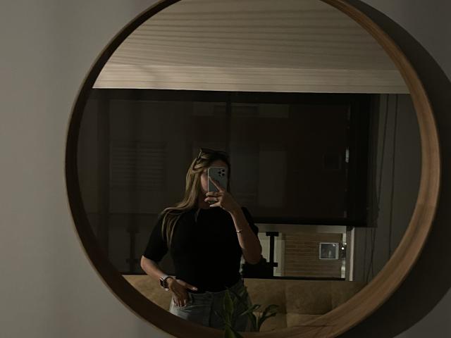 JennyLawry with long blonde hair, black top, and blue jeans, taking a mirror selfie with a wooden circular mirror, holding a phone in front of her face.