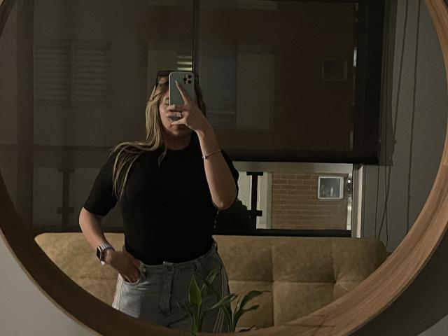 JennyLawry with long blonde hair, wearing a black shirt and light blue jeans, taking a mirror selfie, showing a wrist bracelet, in a dimly lit room.