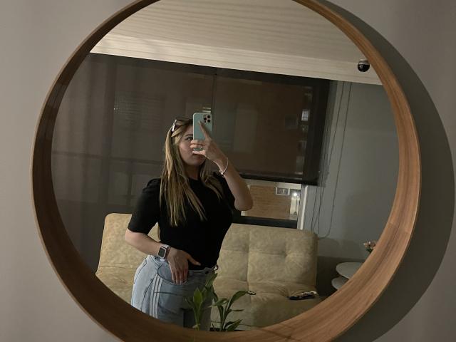 JennyLawry with long blonde hair, black top, and light blue jeans, taking a mirror selfie, showing her ass and a slim waist, holding a green phone.