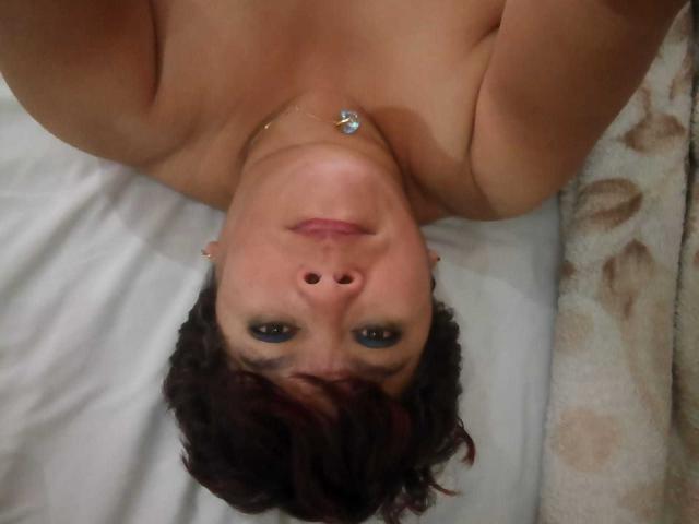 Angellovelly's topless face is upside down in the photo, showing her green eyes, brown wavy hair, and a small silver necklace. Her bare shoulders are visible.