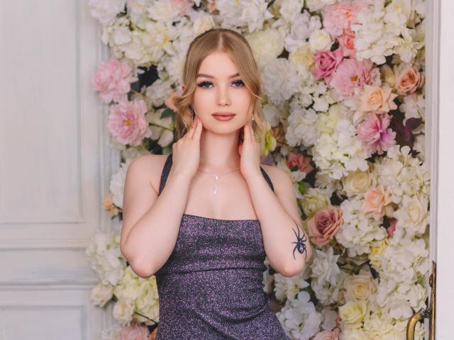 NicoleBrian has blonde hair, blue eyes, wearing a sparkly purple dress, hands on cheeks, small cross necklace, spider tattoo on right arm, standing in front of a floral wall.