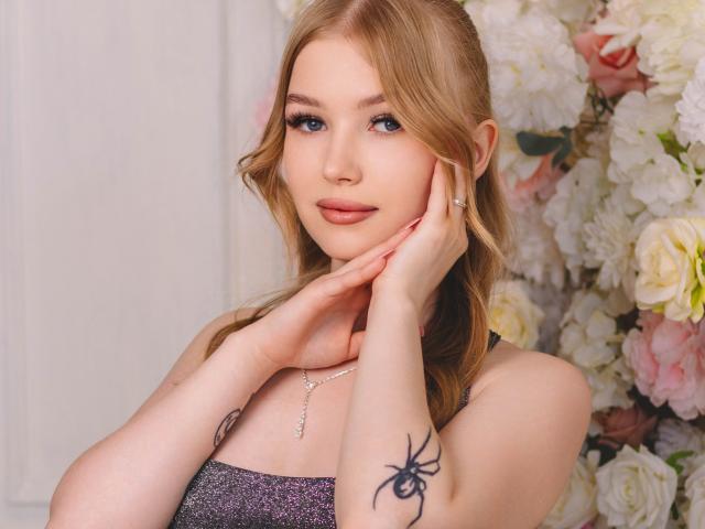 NicoleBrian has blonde hair, blue eyes, and a spider tattoo on her right arm. She's wearing a sparkly top, touching her face with her left hand.