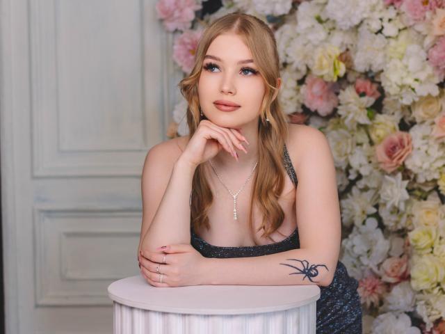 NicoleBrian with blonde hair, blue eyes, and a black spider tattoo on her arm, wearing a sparkly dress, leaning on a white table, looking up with a slight smile.