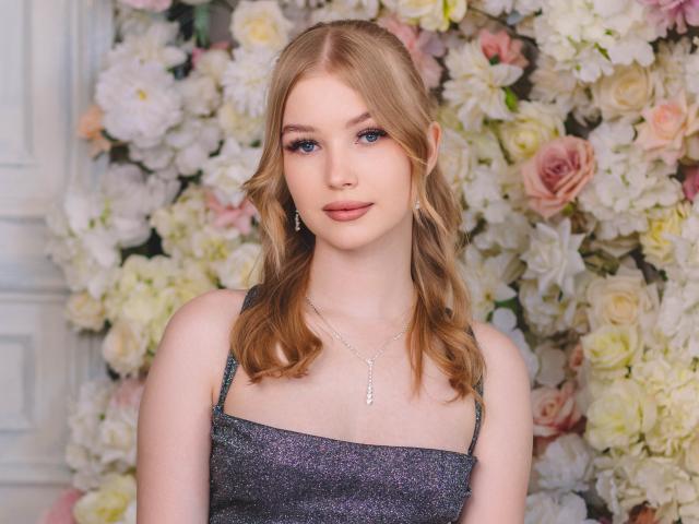 NicoleBrian has blonde wavy hair, blue eyes, and is wearing a sparkly gray dress with a silver necklace. She's got small tits and a cute, sexy face.