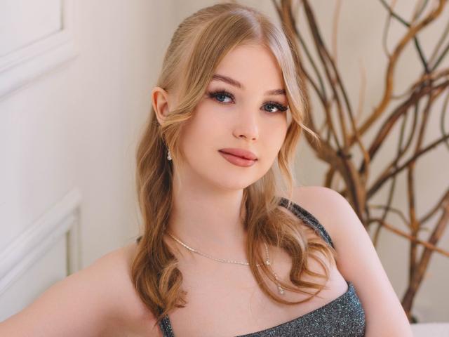 NicoleBrian has blonde pigtails, blue eyes, and pink lipstick. She's wearing a glittery dress with a silver necklace, looking at the camera with a slight smile.